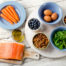 A flat-lay of healthy foods including salmon, carrots, eggs, blueberries, nuts, spinach, dried apricots, and olive oil on a white wooden table.