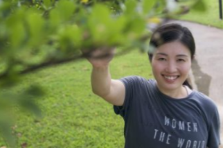 Dr Fiona Wu at outdoors reaching toward a tree branch, wearing a T-shirt with the words “Women of the World.”