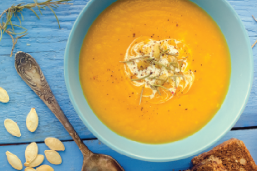 A bowl of spiced pumpkin and cauliflower soup topped with cream, herbs, and cracked pepper, served with toasted bread on a bright blue table.