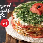 A tall layered vegetable pie stacked with zucchini, pumpkin, sweet potato, mushrooms, and tomatoes, topped with fresh herbs and sliced tomato garnish.