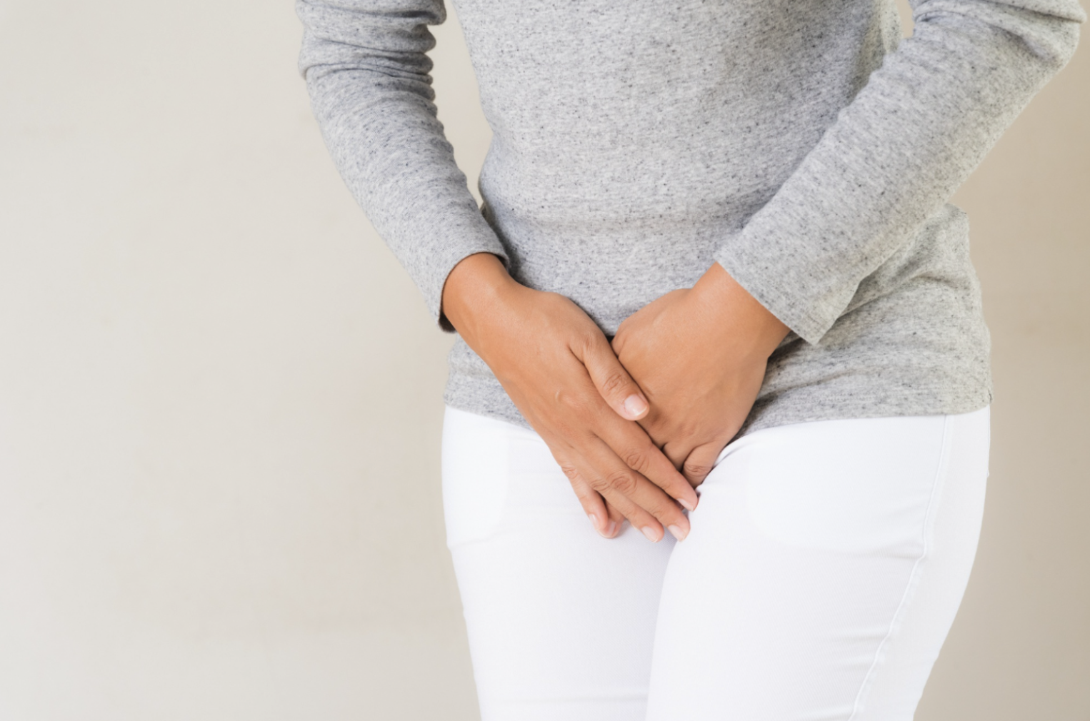 Cropped view of a person in a grey top and white trousers holding both hands over their pelvic area.