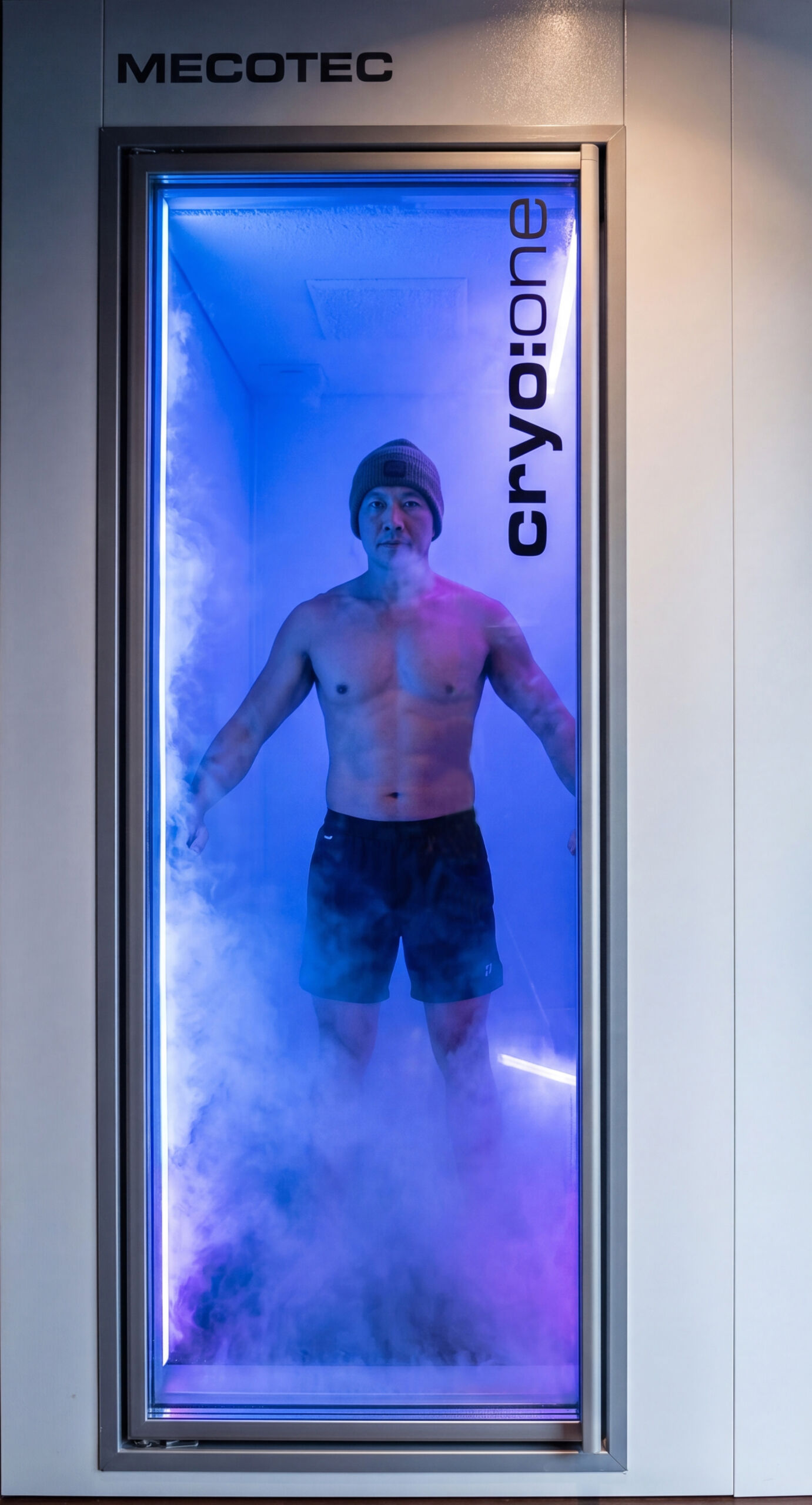Man wearing a beanie and shorts standing inside a blue-lit whole-body cryotherapy chamber at RAPIDÉ.